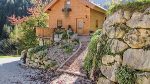 Een houten chalet met een stenen muur, een houten terras en een tuin met planten en een pad.