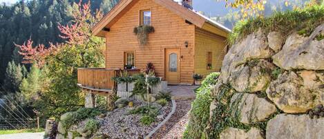 Una cabaña de madera con un muro de piedra, una terraza de madera y un jardín con plantas y un sendero.