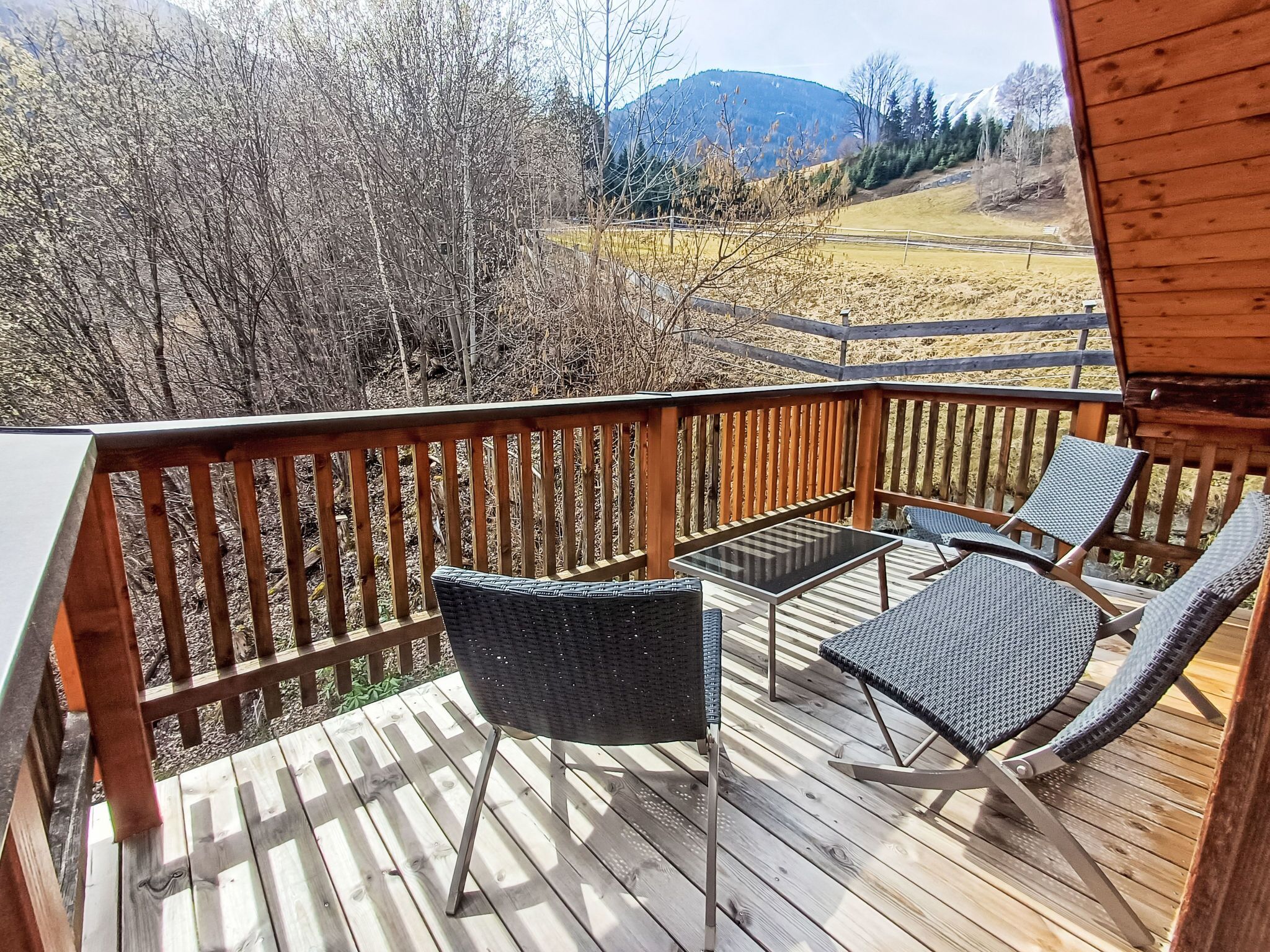 Eine Holzterrasse mit Gartenmöbeln, darunter zwei Stühle und ein Tisch, mit Blick auf eine gebirgige Landschaft.