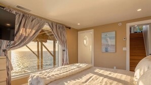 A bedroom with a large bed, a TV, a window with curtains, a painting, and a staircase.