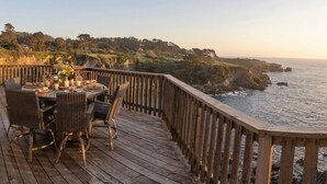 A wooden deck with wicker furniture and a table set for dining, overlooking a coastal landscape with cliffs and the ocean.