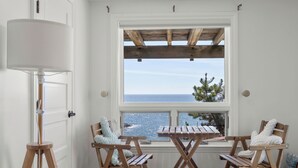A room with a wooden table and chairs, a standing lamp, and a window overlooking the sea.