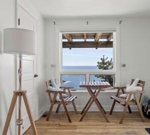 Una habitación con una mesa y sillas de madera, una lámpara de pie y una ventana con vista al mar.
