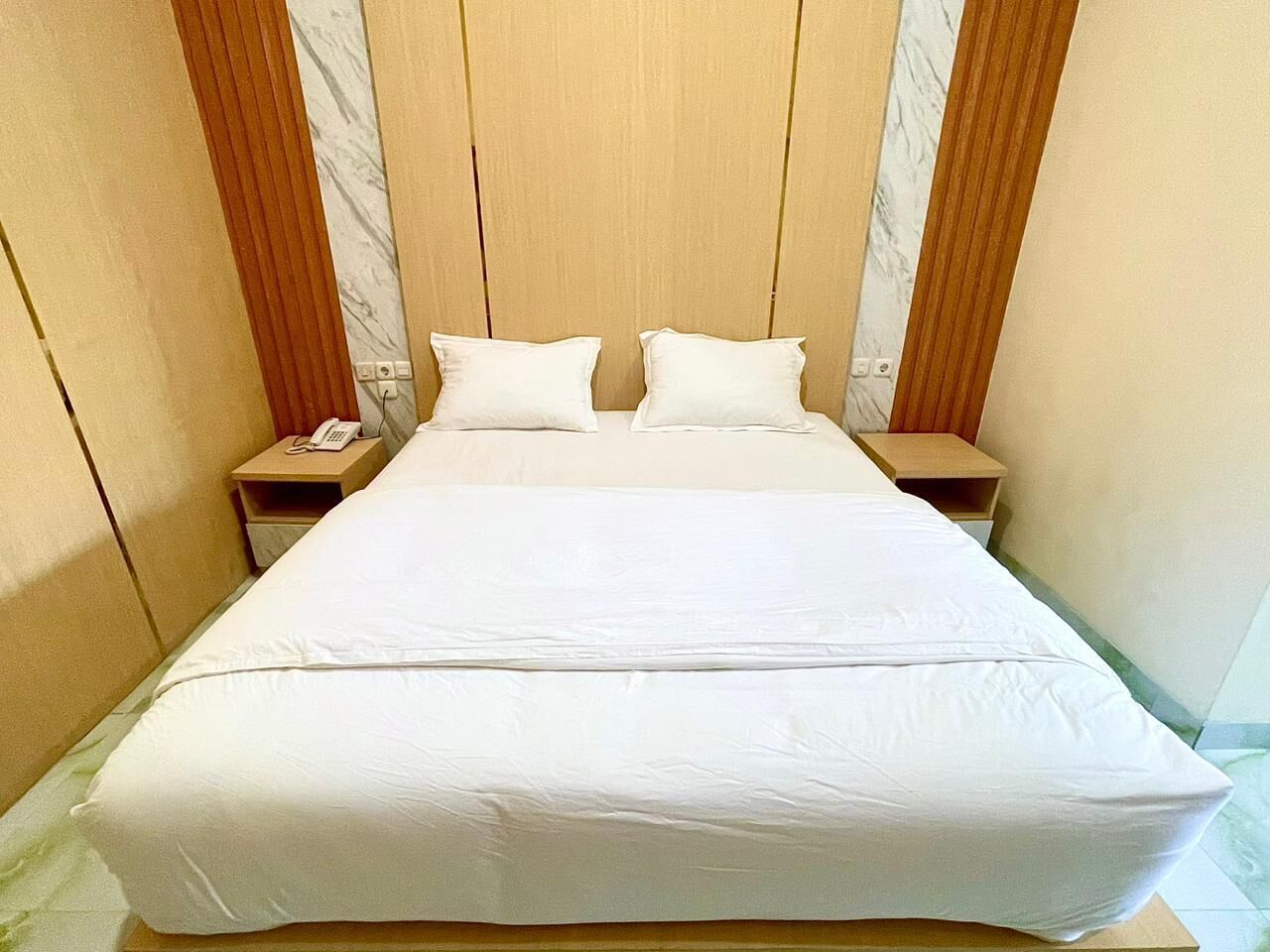 Cama con sábanas blancas, dos almohadas, cabecera de madera y mesitas de noche con un teléfono.