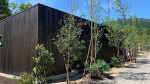 Un edificio moderno con fachada de madera oscura, rodeado de árboles jóvenes y un cielo despejado.