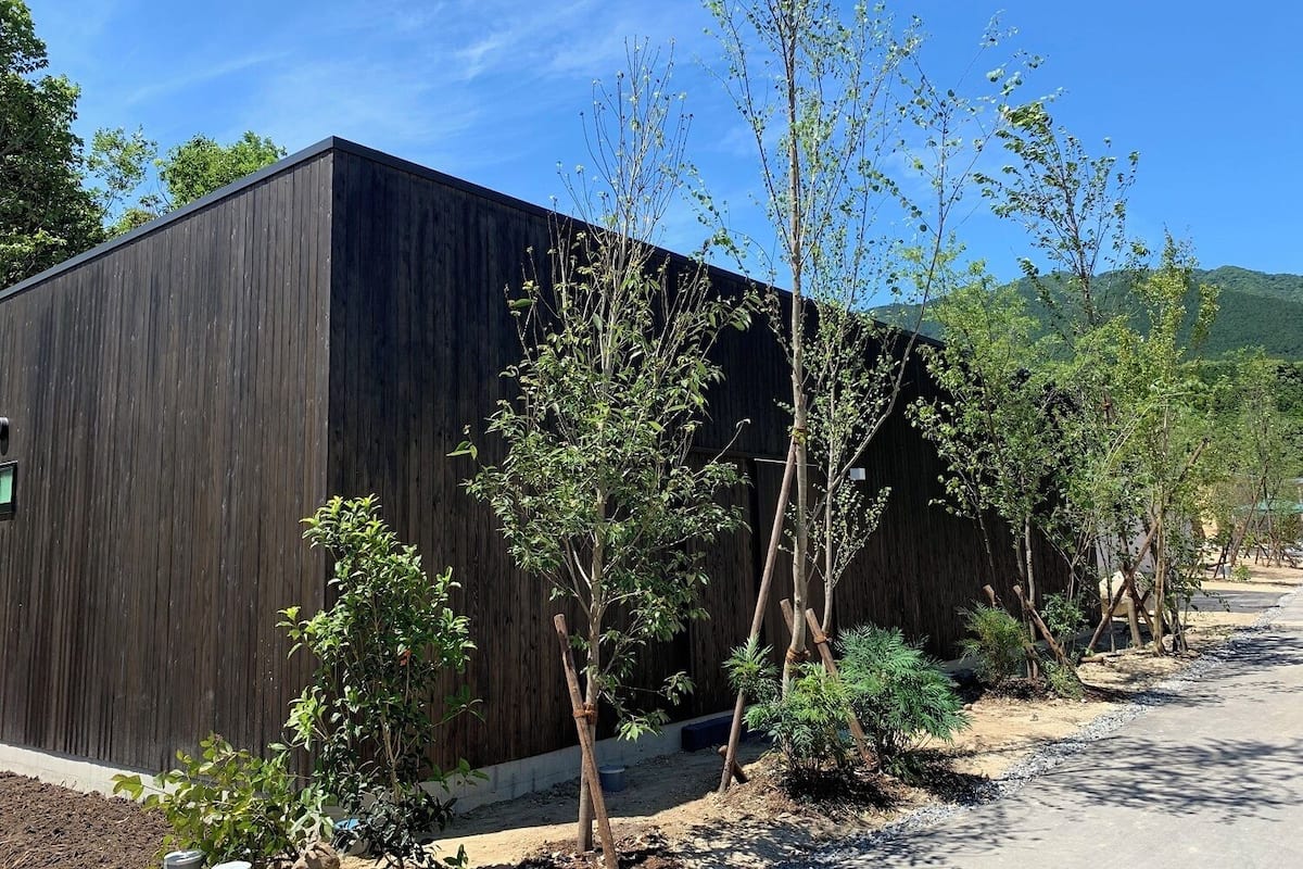 A modern building with a dark wooden exterior, surrounded by young trees and a clear sky.