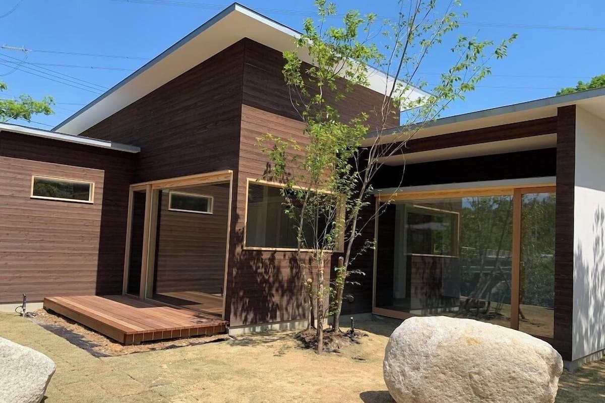 A modern house with a wooden exterior, large windows, a small tree, and a rock.