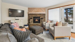 A living room with a stone fireplace, a flat-screen TV, a sofa, a chair, and a large window. - New Listing! Whitefish Lake Condo, 16B, Hillside! (Whitefish)