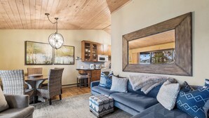 A cozy living room with a wooden ceiling, a dining area with wicker chairs, a blue sofa with patterned cushions, and a kitchen with wooden cabinets. - New Listing! Whitefish Lake Condo, 16B, Hillside! (Whitefish)