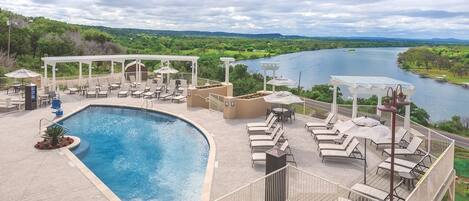 A pool area with lounge chairs and umbrellas, surrounded by a deck with a view of a river and hills.