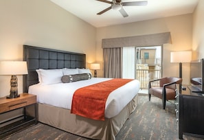 A hotel room with a bed, two bedside tables, a desk with a chair, a ceiling fan, and a view of the outside.