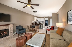 A living room with a fireplace, a ceiling fan, a TV, a dining area with chairs, and a kitchen in the background. - Marble Falls | Group Lake Getaway | Pool + Lake | 3BR Sleeps 10 (Marble Falls)