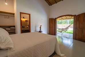 Ein Schlafzimmer mit Bett, Spiegel, Nachttisch, Lampe und Blick auf eine Hängematte im Freien.