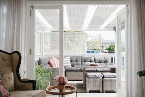 A covered patio with wicker furniture, a cushioned sofa, and a small table with a vase. - Charming Howick Home with Sunny Covered Deck | Marigold by Homello (Howick)