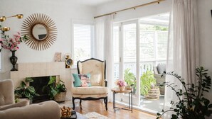 Una sala de estar con chimenea, un sillón con cojín, una mesa redonda y un amplio ventanal con cortinas.