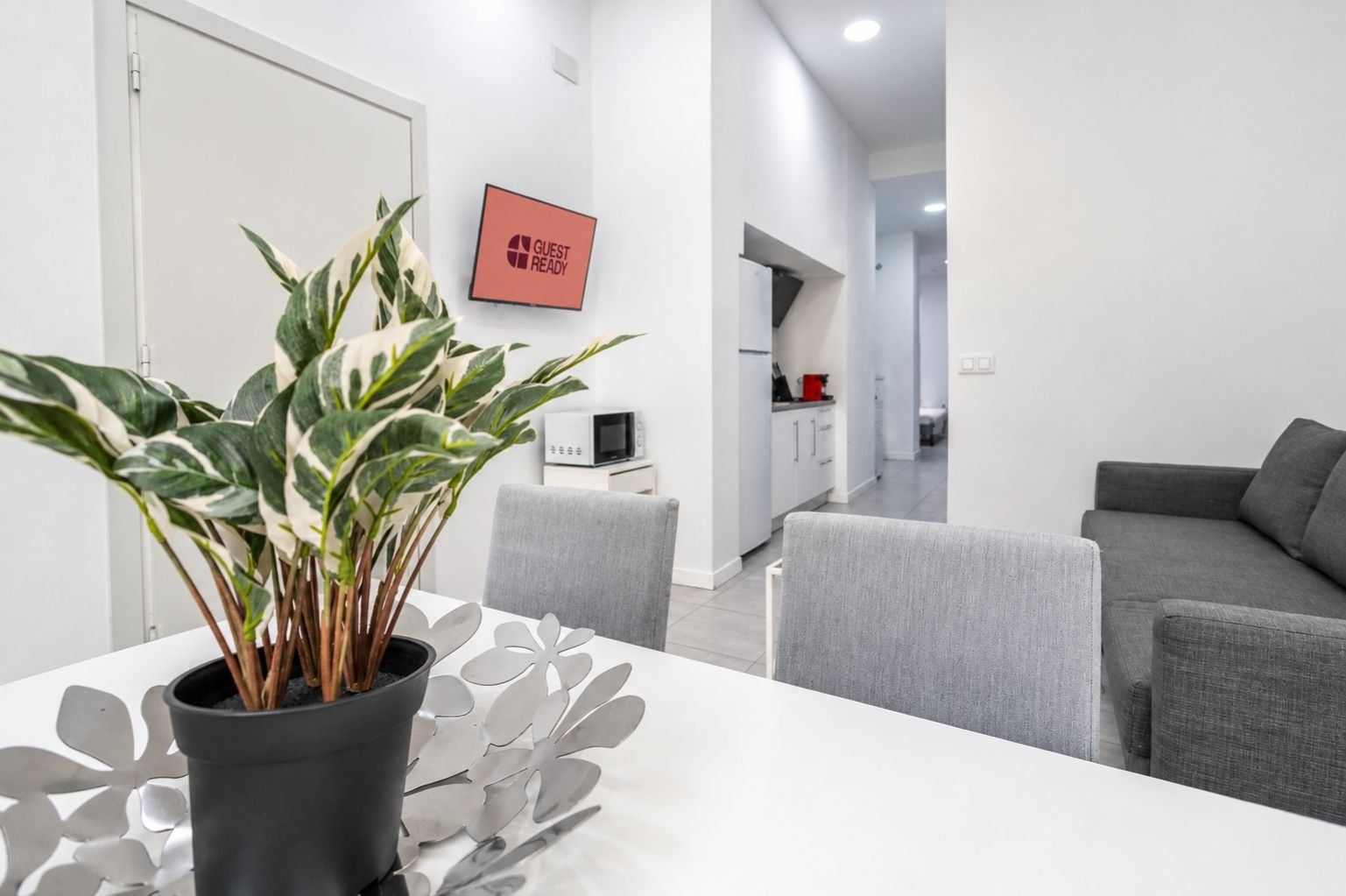 A modern living room with a grey sofa, a dining table with a potted plant, and a kitchen in the background.