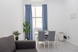 Ein modernes Zimmer mit grauem Sofa, einem Esstisch mit Stühlen, einem Blumentopf und einem Fenster mit Vorhängen.