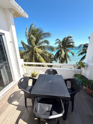Un balcón con mesa y sillas, con vista a una playa con palmeras y el océano.