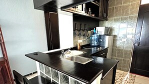 Una cocina compacta con encimera negra, un lavamanos y una estufa. Hay armarios sobre la encimera y un dispensador de agua grande.