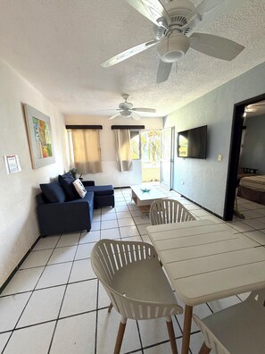 Una sala de estar con ventilador de techo, televisión, sofá, mesa de comedor con sillas y vistas al exterior a través de una ventana.