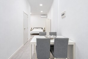 Habitació d'hotel moderna amb zona de menjador que inclou una taula blanca i cadires grises, un llit amb roba de llit blanca i un aire condicionat fixat a la paret.