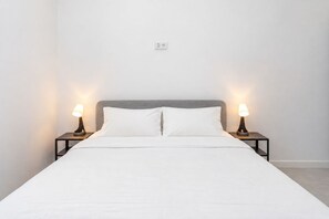 Un llit amb roba de llit blanca, dues taules de nit amb lampades i una paret sense adorns amb una presa elèctrica.