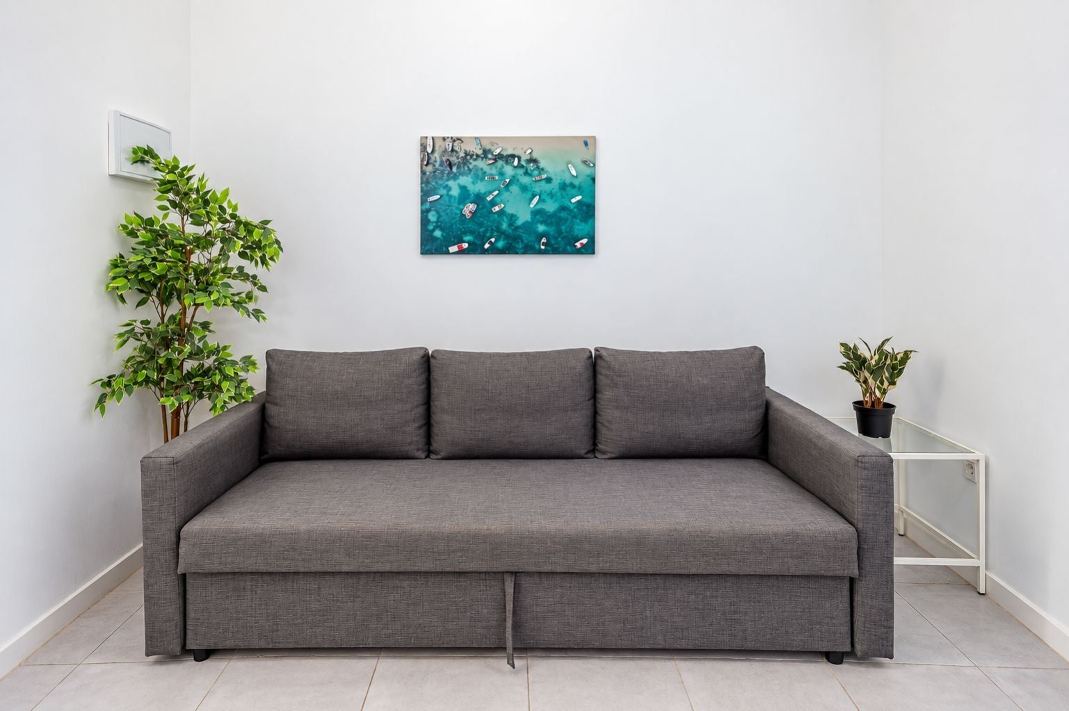 Un sofá gris con respaldo bajo, un cuadro colgado en la pared, una planta en maceta y una mesita pequeña.
