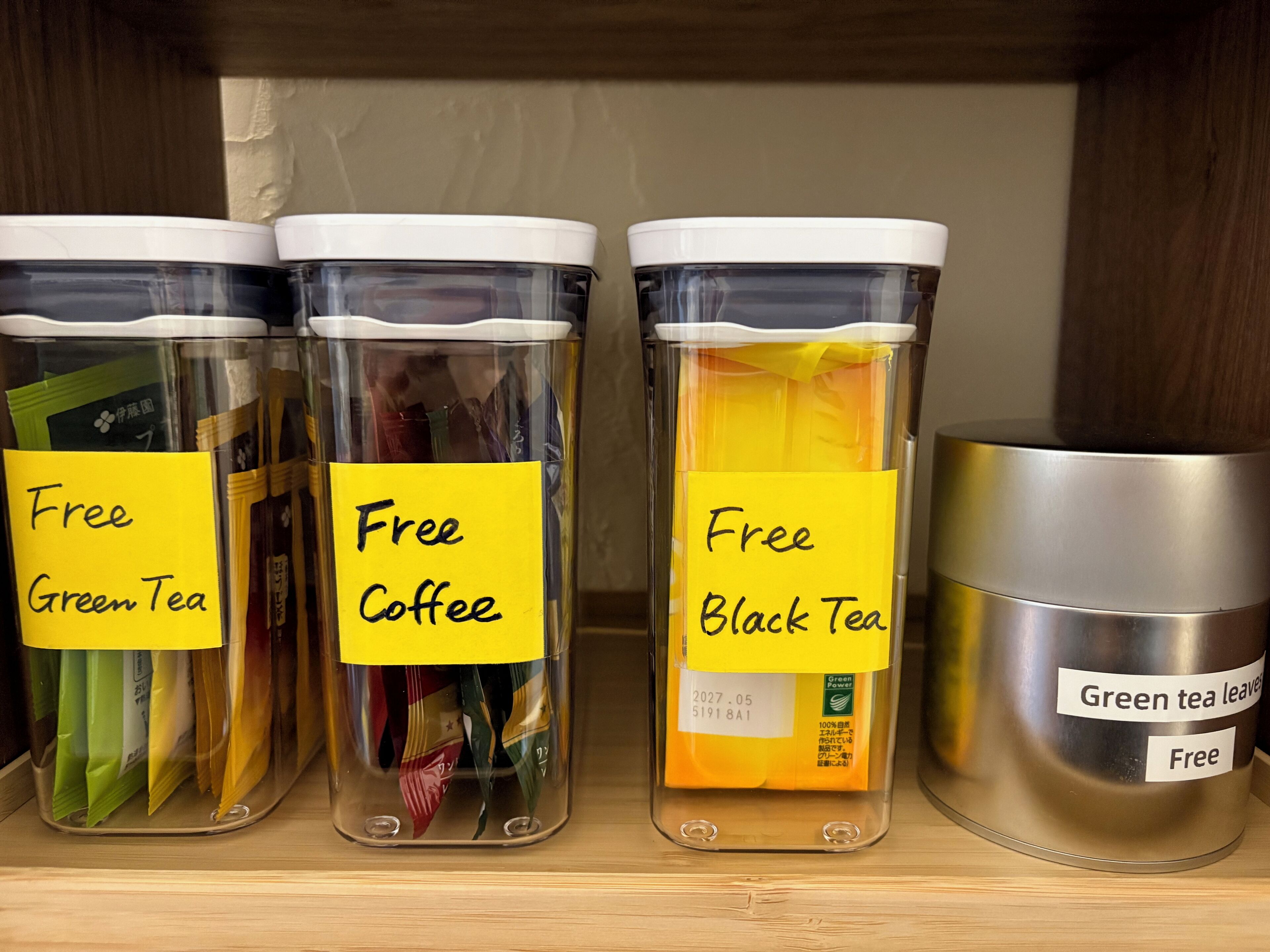 Three transparent containers labeled 'Free Green Tea,' 'Free Coffee,' and 'Free Black Tea,' with a stainless steel container labeled 'Green Tea Leaves' on a shelf.