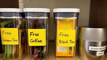 Three transparent containers labeled 'Free Green Tea,' 'Free Coffee,' and 'Free Black Tea,' with a stainless steel container labeled 'Green Tea Leaves' on a shelf.