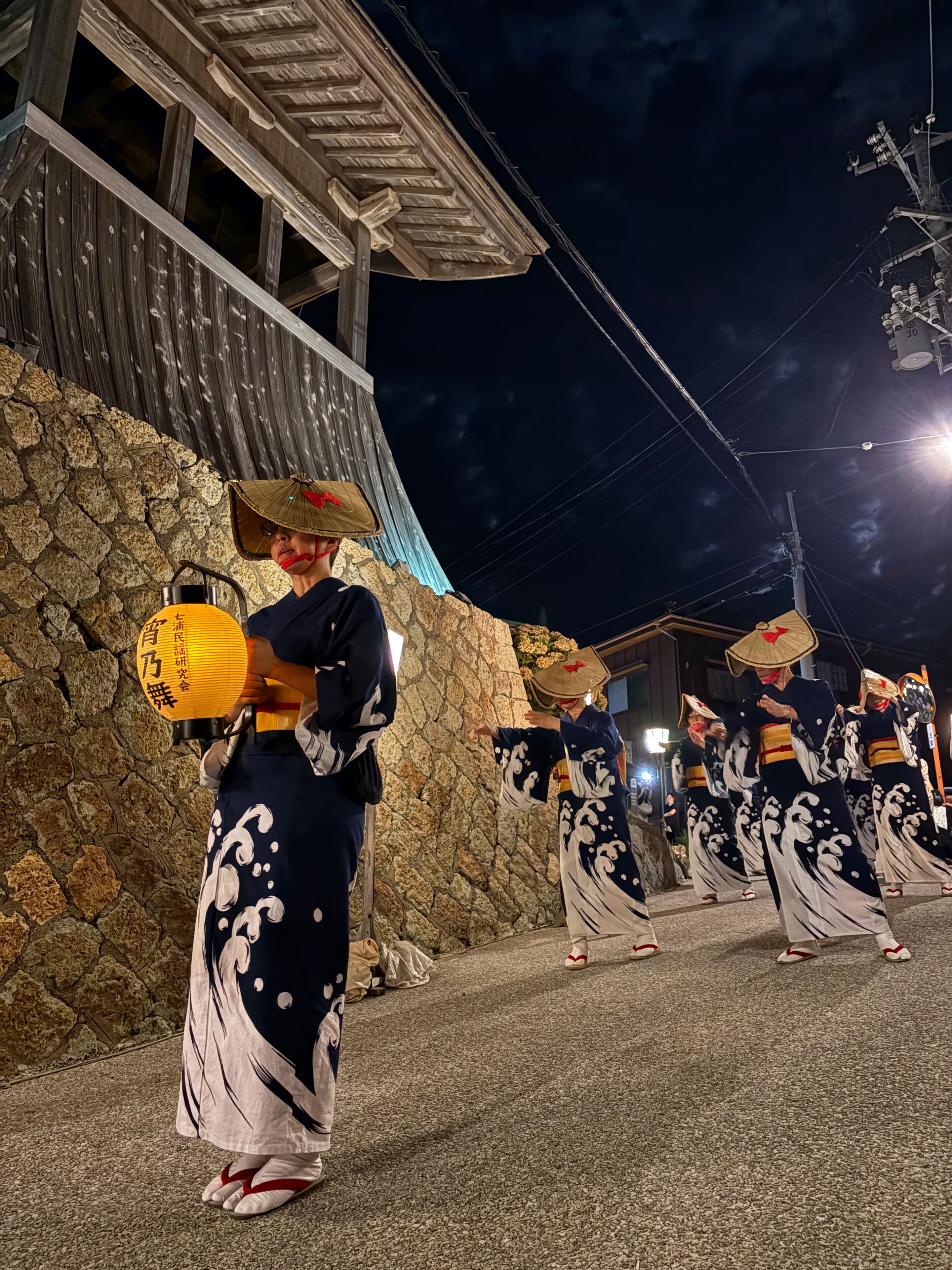 A traditional Japanese festival with performers in kimonos and conical hats, a stone wall, and a wooden building with a sloped roof.