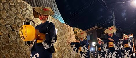 A traditional Japanese festival with performers in kimonos and conical hats, a stone wall, and a wooden building with a sloped roof.