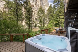 A hot tub on a wooden deck with a view of a rocky cliff and trees. - Experience a riverfront Big Sky home with hot tub! (Gallatin Gateway)