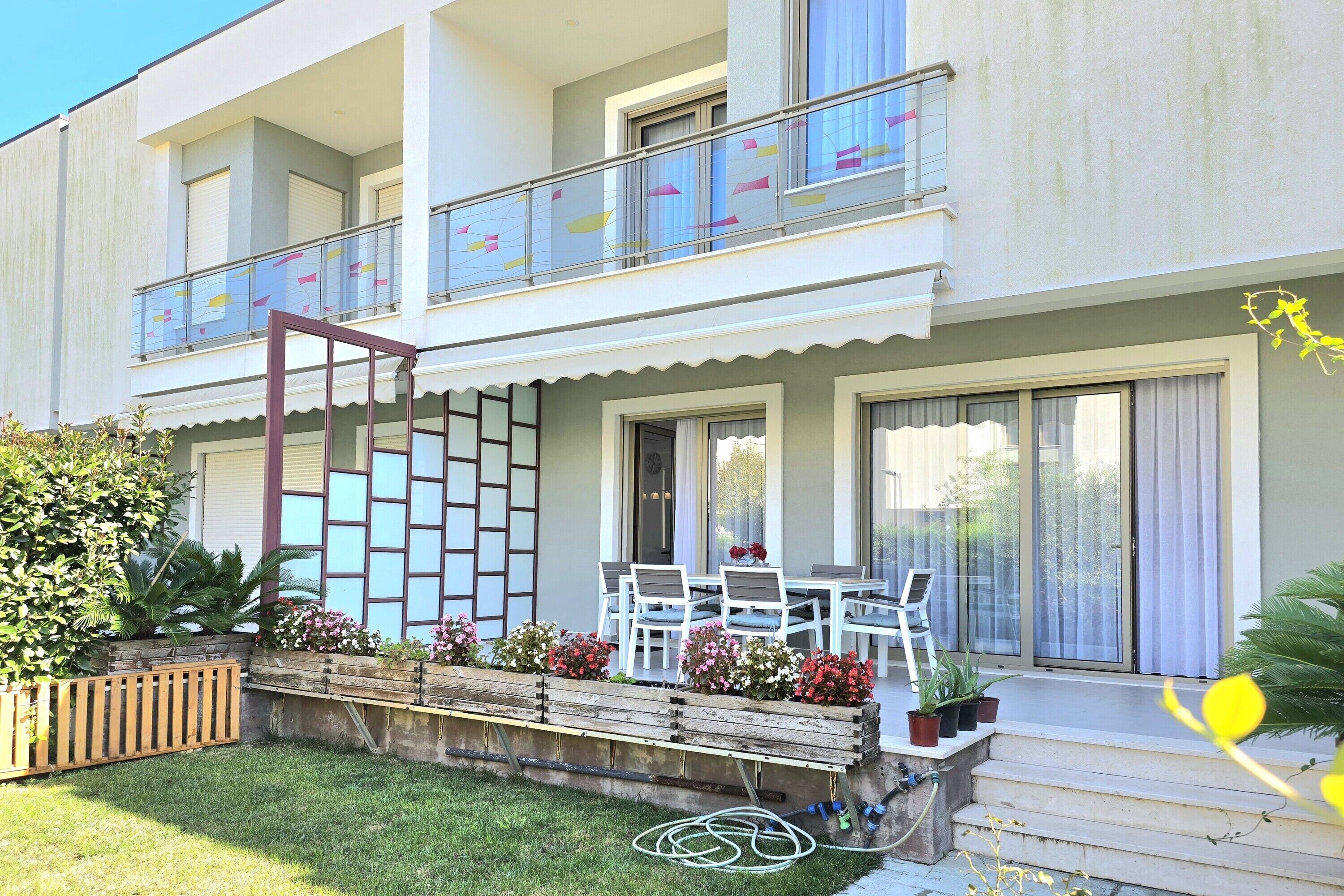 Un bâtiment à deux étages doté d’un balcon, d’un petit espace extérieur aménagé avec des chaises et une table, et d’un jardin avec des plantes et des fleurs en pot.