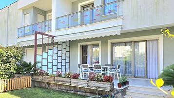 Un bâtiment à deux étages doté d’un balcon, d’un petit espace extérieur aménagé avec des chaises et une table, et d’un jardin avec des plantes et des fleurs en pot.