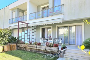 Un bâtiment à deux étages doté d’un balcon, d’un petit espace extérieur aménagé avec des chaises et une table, et d’un jardin avec des plantes et des fleurs en pot.