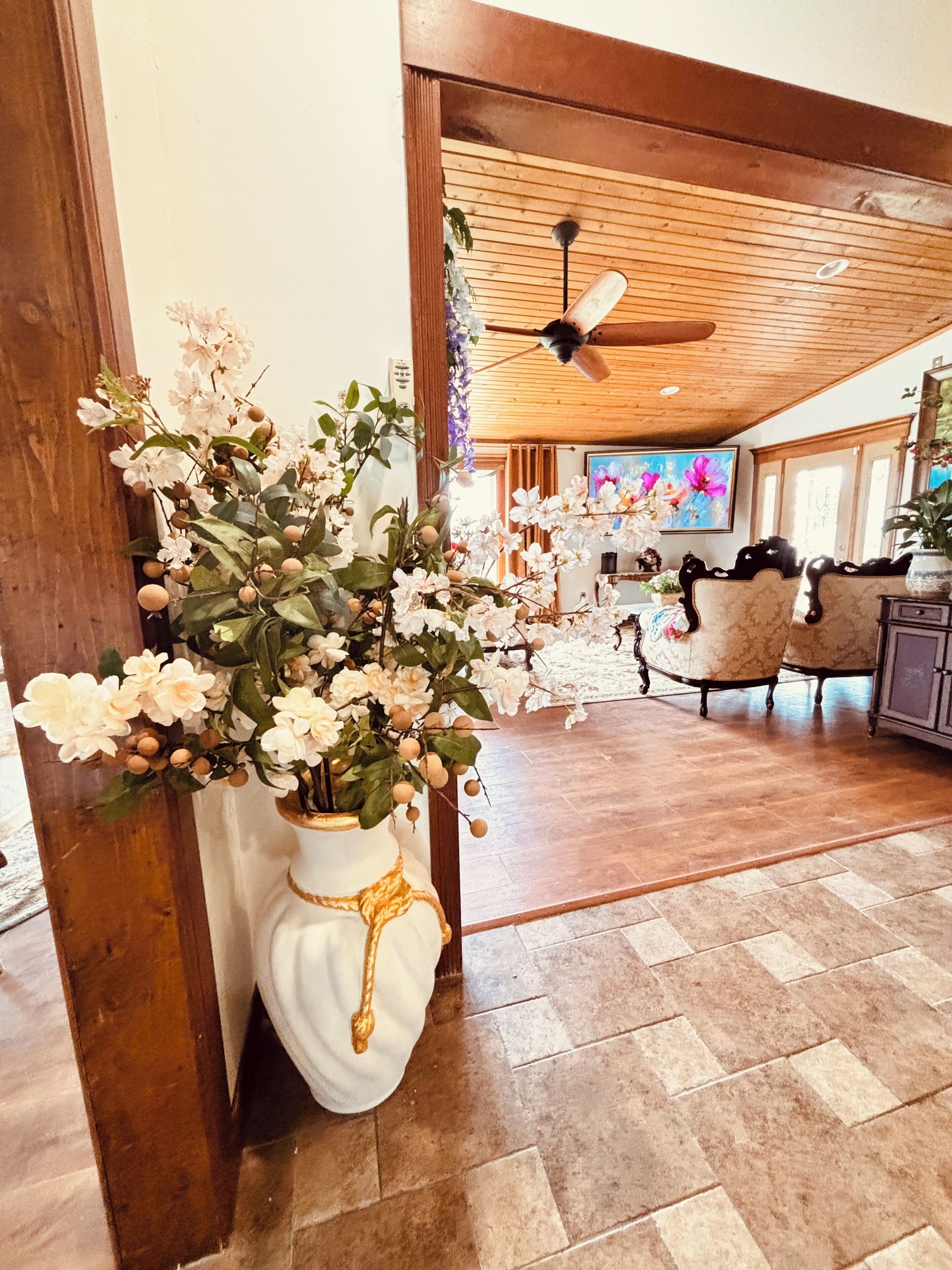 A room with a wooden ceiling, a ceiling fan, a white vase with flowers, and a patterned floor.