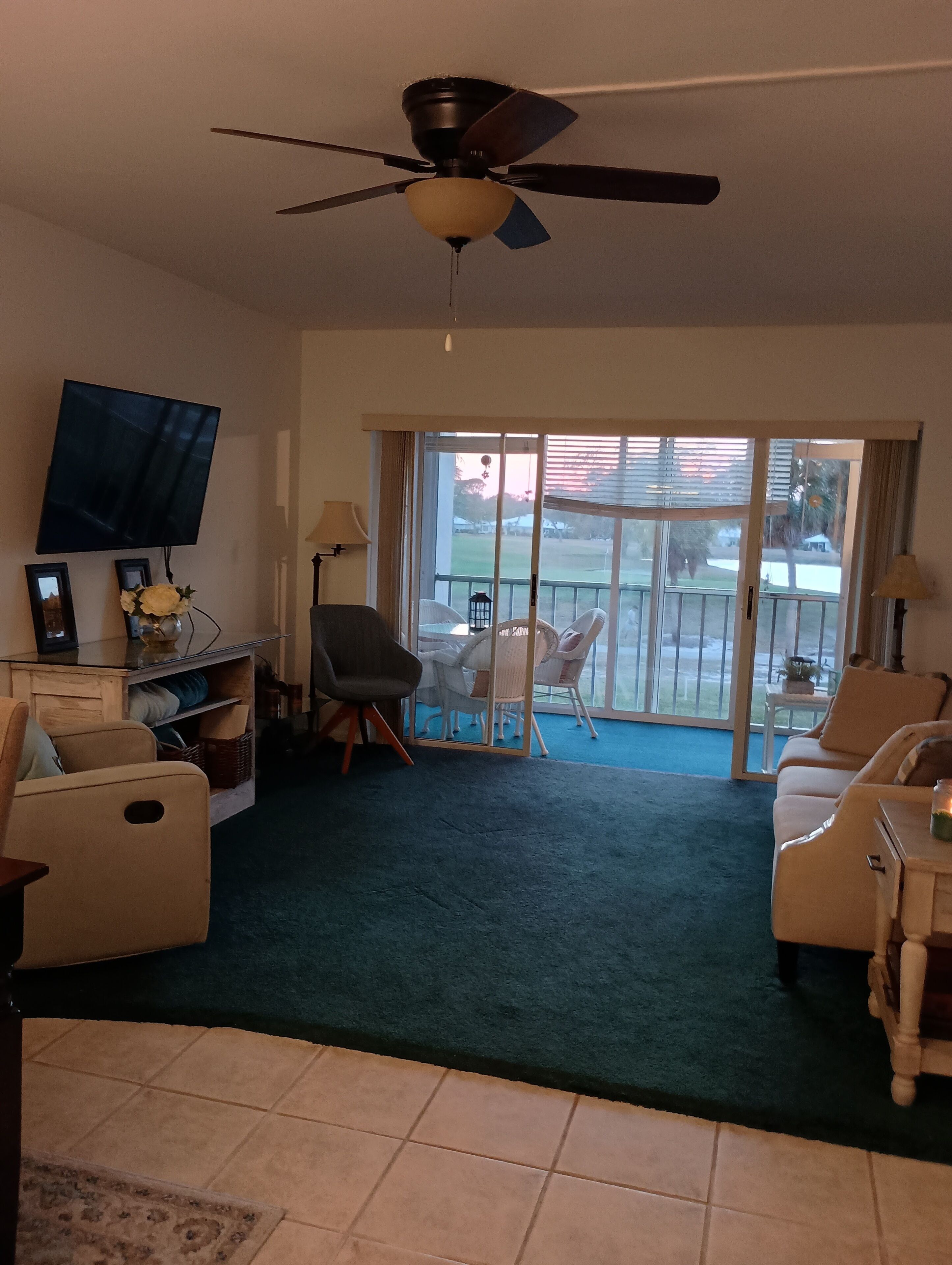 A living room with a ceiling fan, a flat-screen TV, a sofa, a chair, a small table, and a view through a sliding glass door to an outdoor area with a patio.
