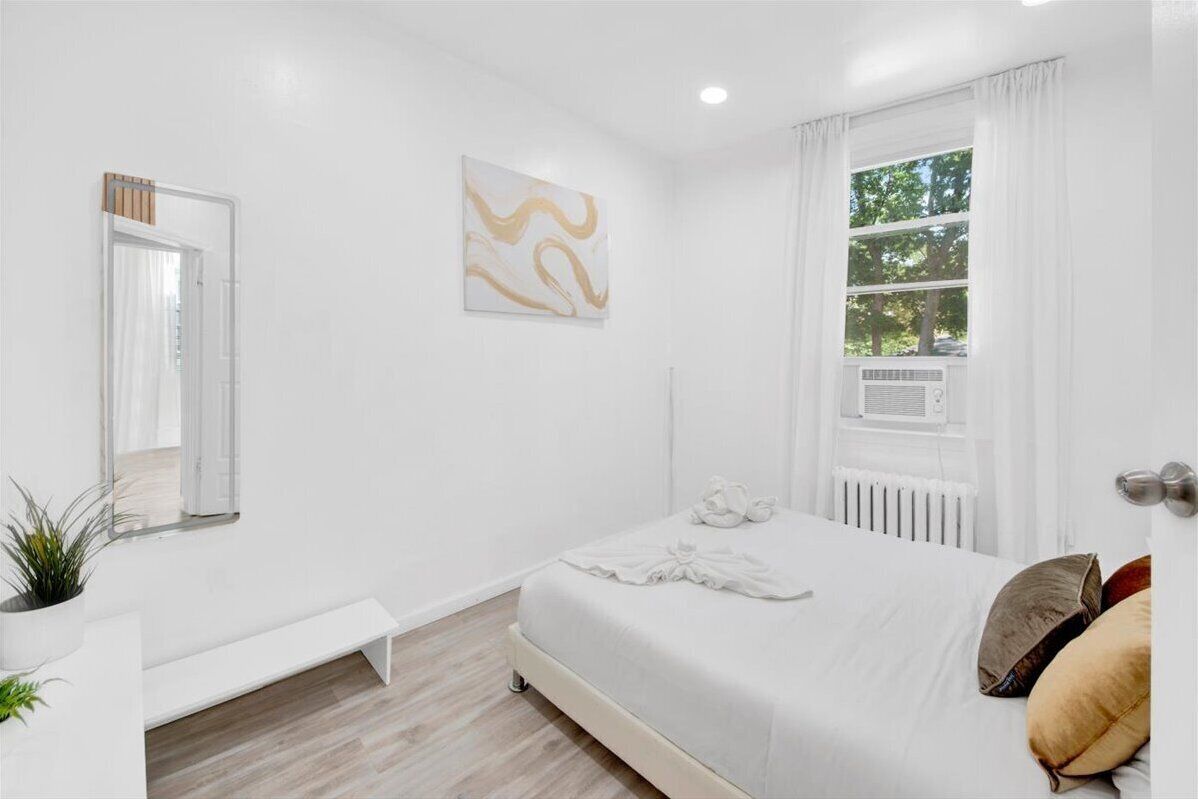 Une chambre à coucher moderne, avec un lit, une table de chevet, un banc, un radiateur et une fenêtre avec des rideaux.