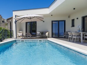A modern outdoor pool area with a clear blue pool, white lounge chairs, a table with chairs, and a large umbrella. - Florinda by Interhome (Rabac/Sveti Lovreč Labinski)