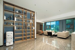 A modern living room with a large bookshelf, a sofa, a coffee table, and a large window with a view of greenery. - HotelYizhiStationemMetroAurum  (Guangzhou)