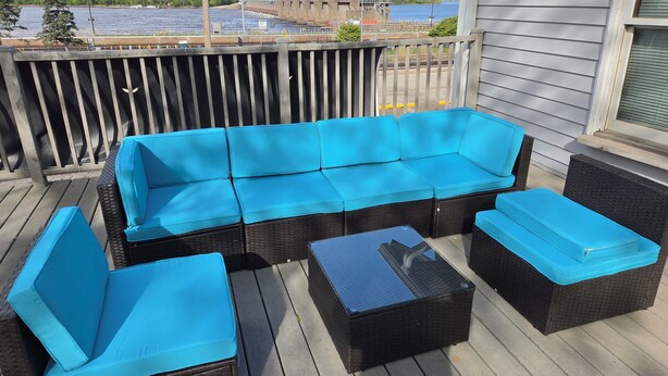 Een overdekt terras met een bank en stoelen, een kleine tafel en uitzicht op een waterpartij en industriële gebouwen op de achtergrond.