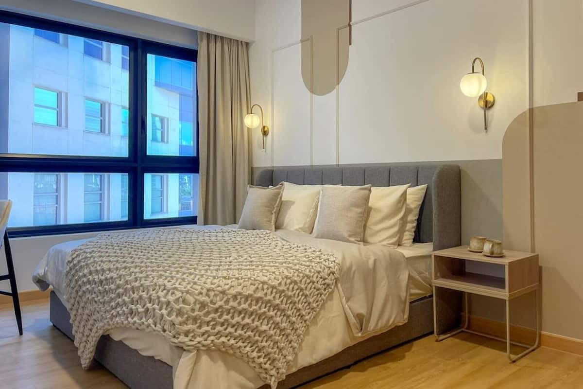 A modern bedroom with a large bed, a bedside table, a wall-mounted light, and a window with curtains.