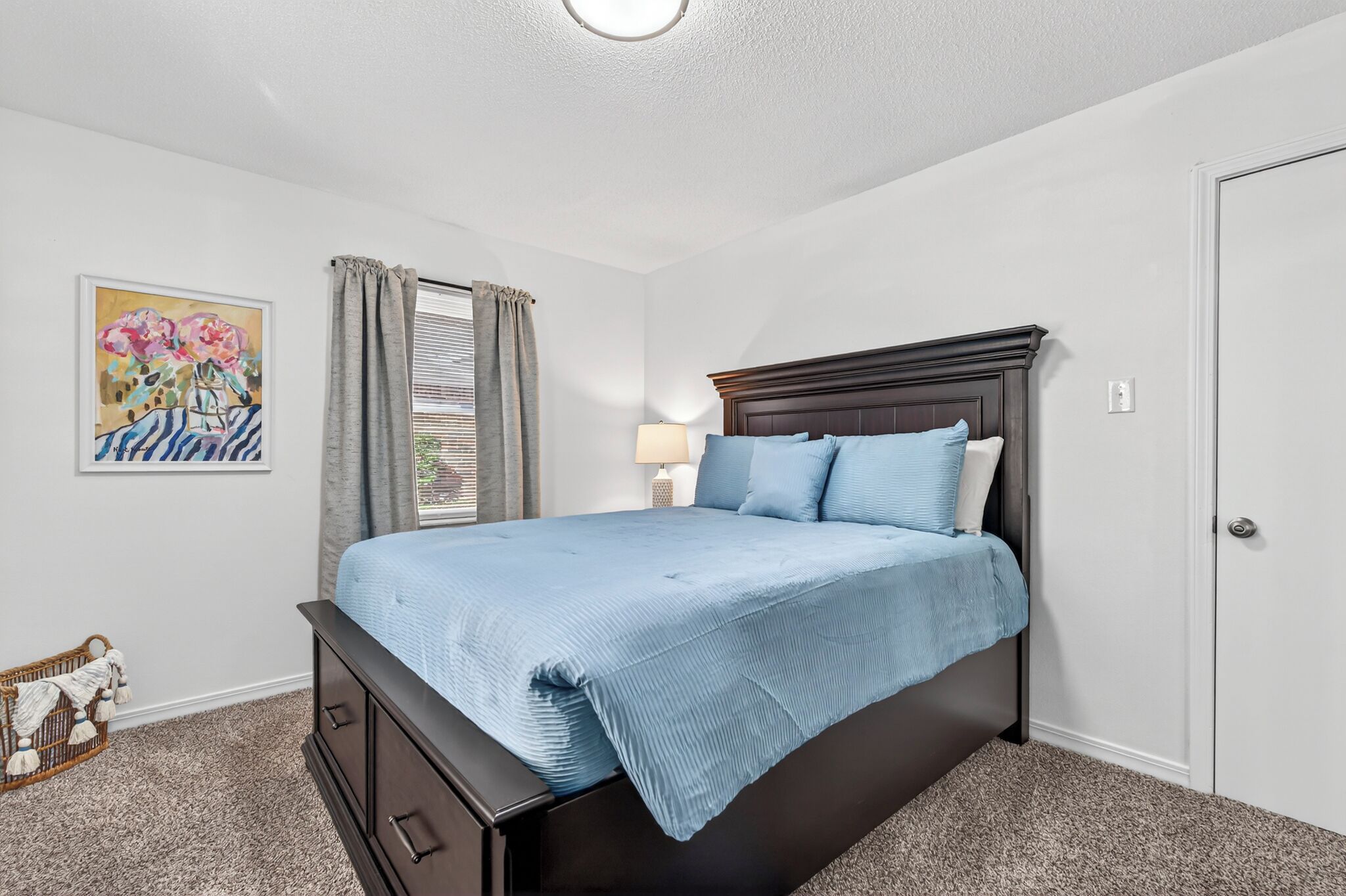 A bedroom with a bed, a nightstand, a lamp, a painting, and a door.