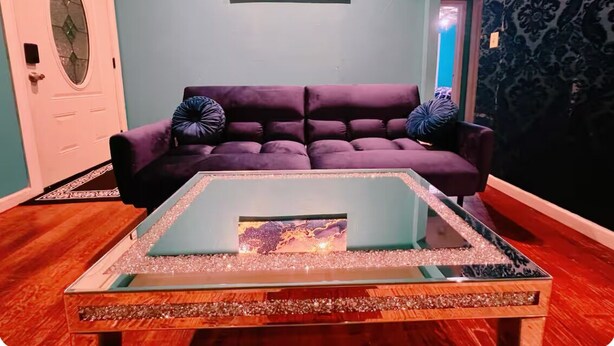 En stue med en lilla sofa, et sofabord af glas med et marmorlignende mønster, og en væg med tapet.