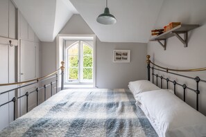 A bedroom with a checkered bedspread, a window with a view of greenery, a wall-mounted shelf with books, and a hanging light fixture. - Daphne Cottage, Orford, Suffolk (Orford)