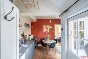 A modern kitchen with a dining area, a red accent wall, and a view through a large window. - Daphne Cottage, Orford, Suffolk (Orford)