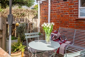 A patio with a round table, a bench, and a potted plant with white flowers. - Daphne Cottage, Orford, Suffolk (Orford)