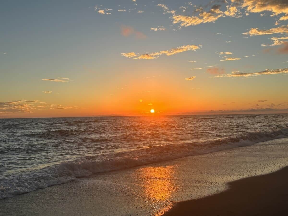 Ein Strand mit untergehender Sonne über dem Meer, sanfte Wellen schlappen an das Ufer.