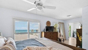 Ein Schlafzimmer mit einem Bett, einem Deckenventilator, einem Fernseher, einer Kommode und Blick auf den Ozean durch große Fenster.