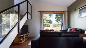 Una sala de estar con un sofá de cuero negro, un televisor sobre un mueble bajo, una escalera y un ventanal con cortinas.